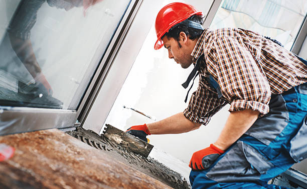 Closeup side view of a handyman installing ceramic tiles on the floor. He's applying some tile glue with a trowel before laying the tiles. The worker is wearing blue uniform, red work helmet and gloves.